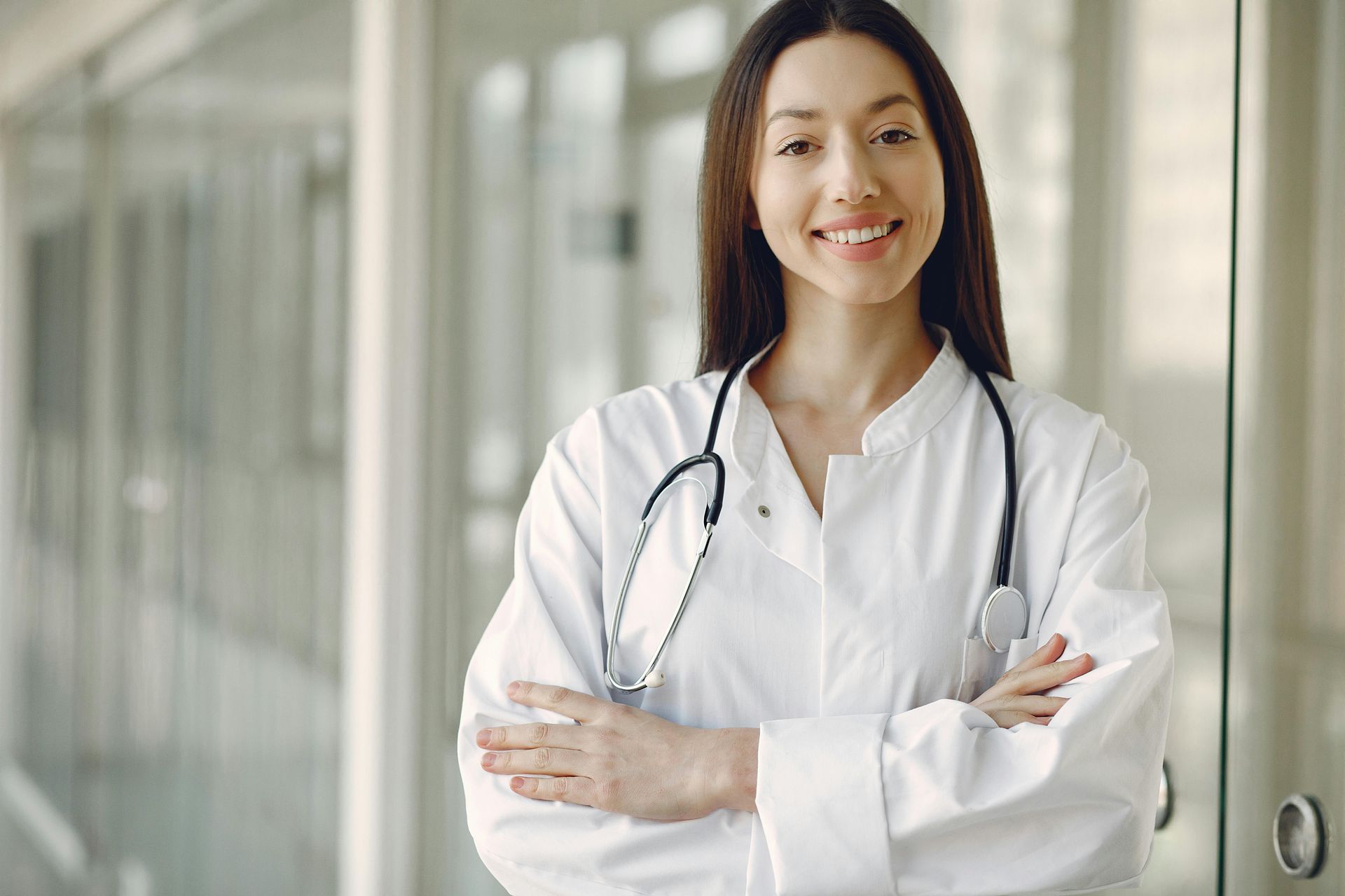 Smiling female doctor with crossed arms, wearing a white coat and stethoscope, standing in a hallway.