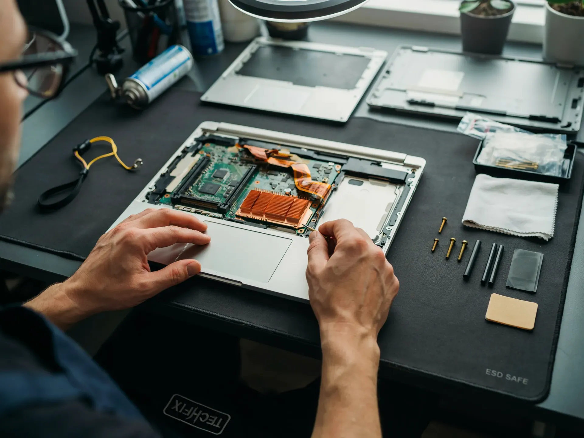 Hands repairing a laptop motherboard on a workbench with tools and small parts.