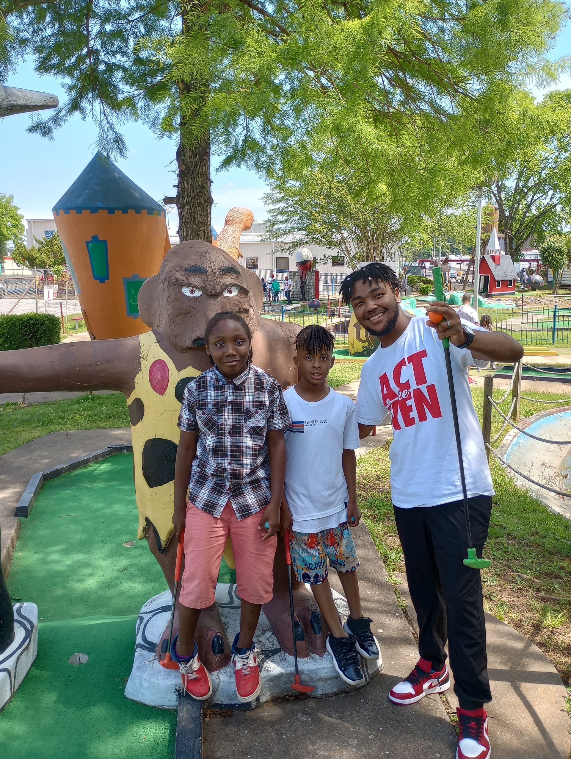 Three people posing near a miniature golf course statue. A person in a white shirt holds a golf club. Two children stand next to the statue.