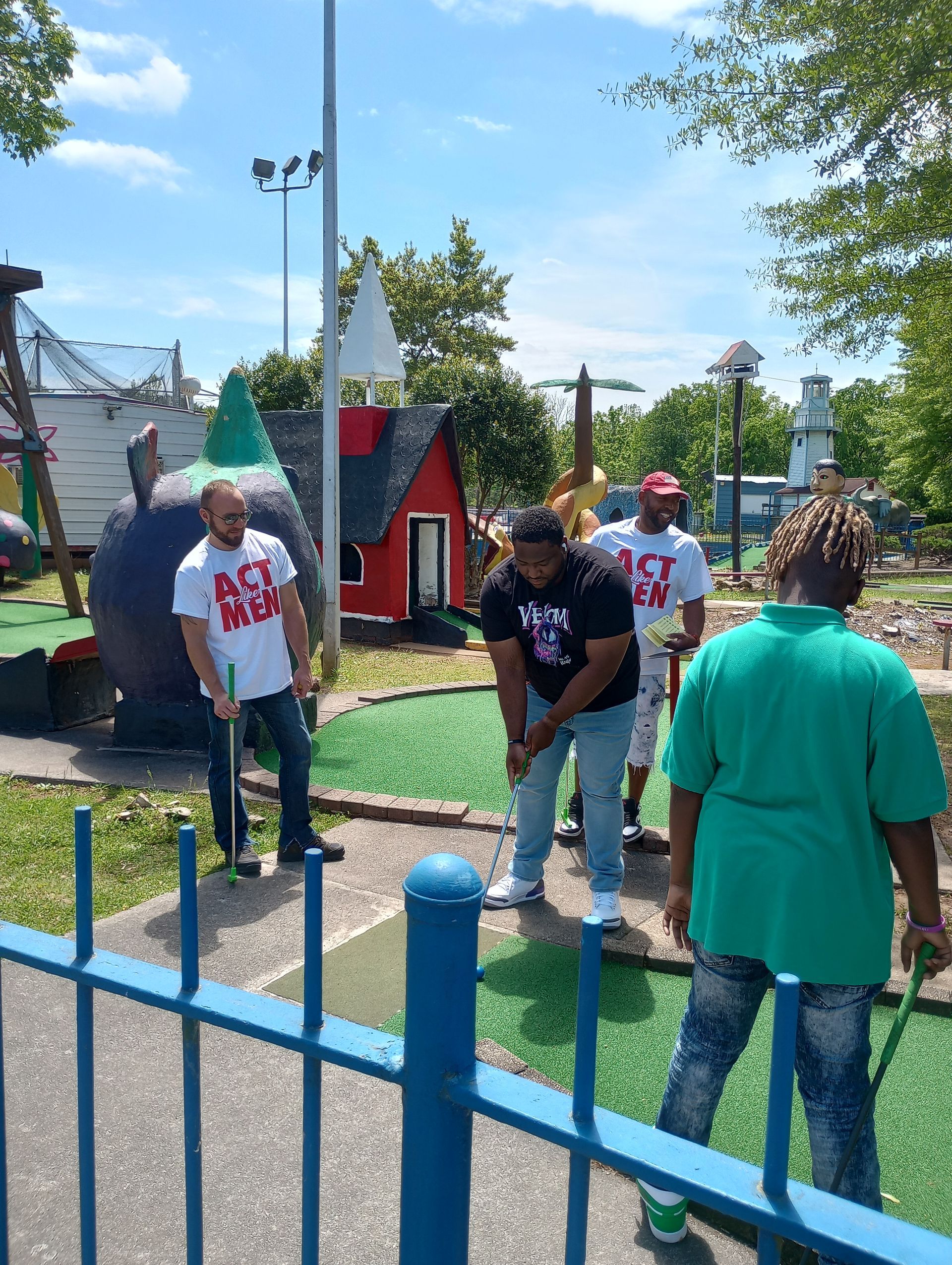 Group of people playing mini golf on a sunny day. Some are wearing white t-shirts with 