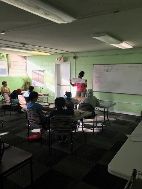 A teacher presents a document to students in a classroom. Students sit at desks, facing forward. The room is painted light green.