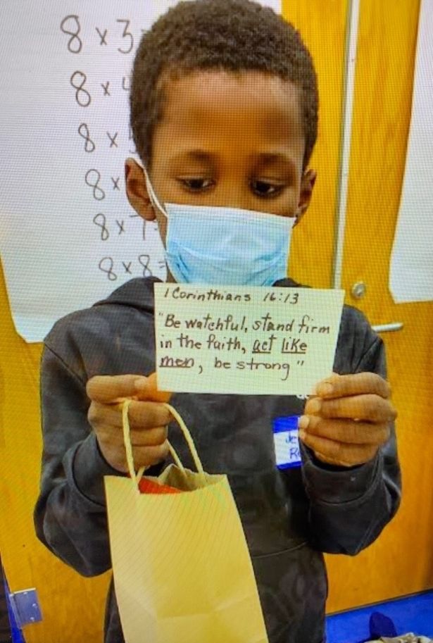 A young boy wearing a mask holds a card with a Bible verse and a gift bag in a classroom setting. He appears focused and earnest.