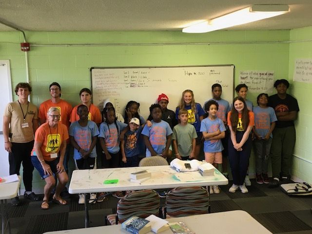 Group of kids and adults pose in a classroom, in front of a whiteboard. Most wear matching blue shirts.