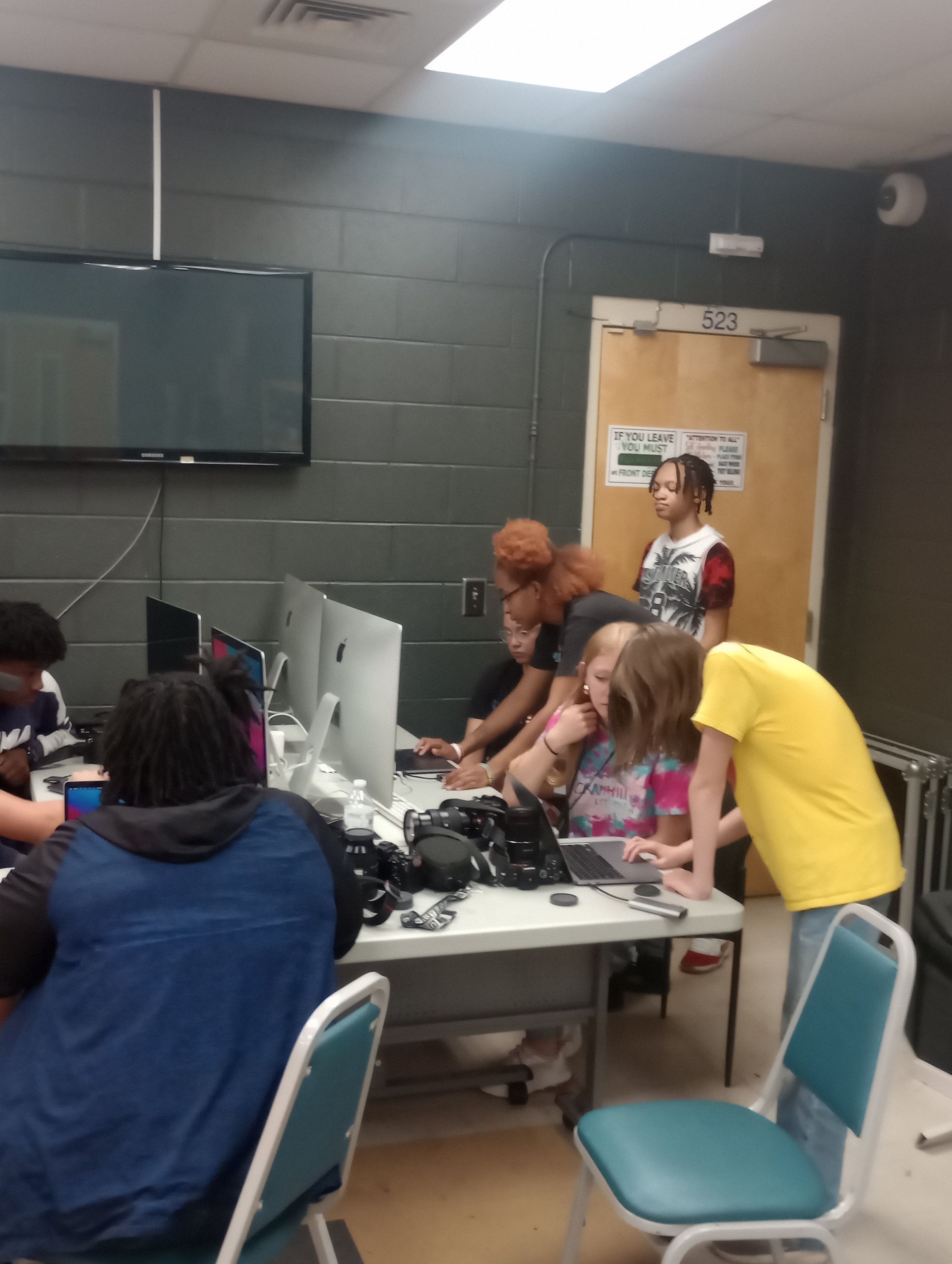 A group of students huddled around a computer in a classroom, focused on the screen. One student wears a yellow shirt.