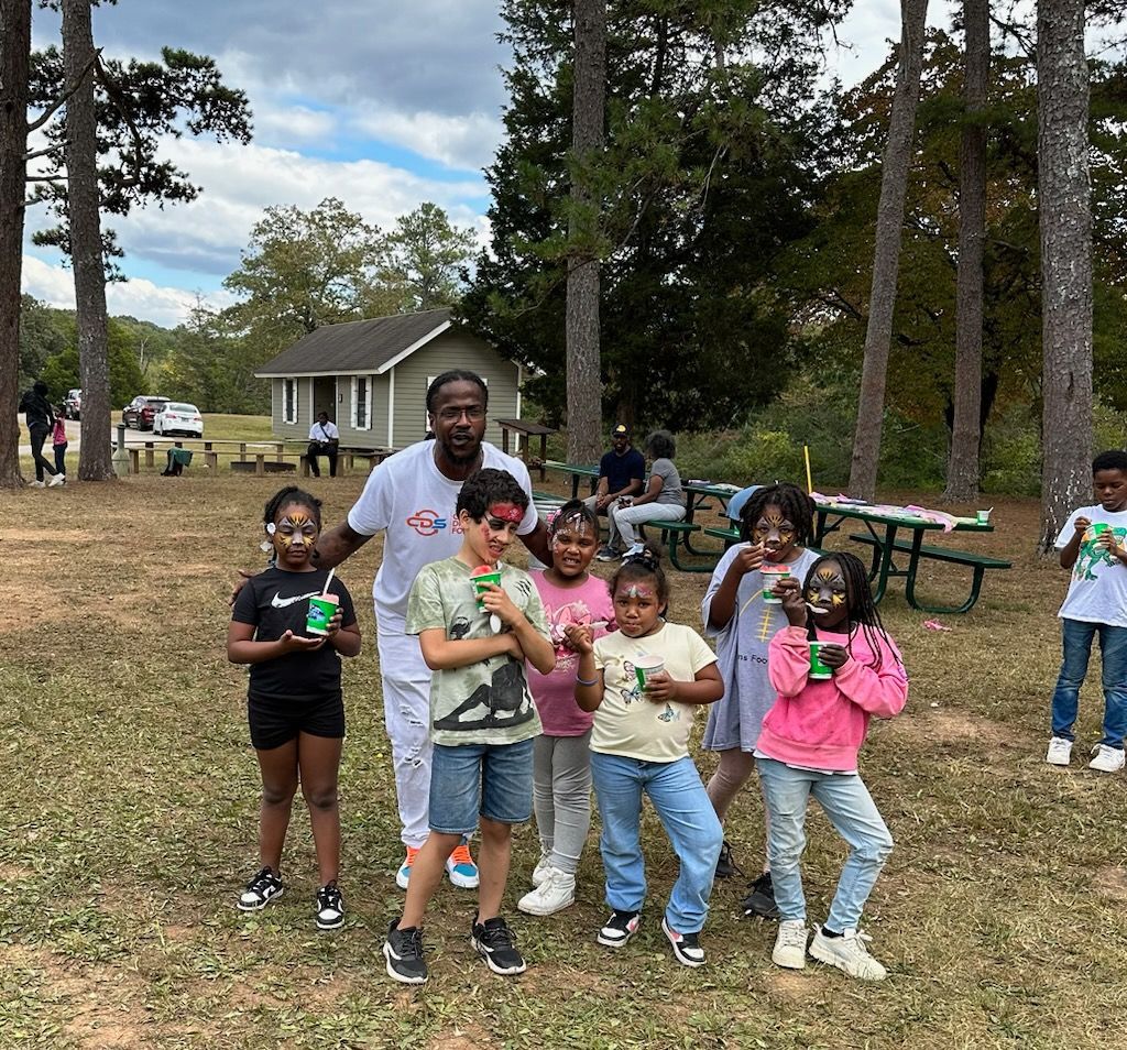 Group of kids and a man in a park, some with face paint, holding drinks. They are smiling outdoors near trees and a small building.