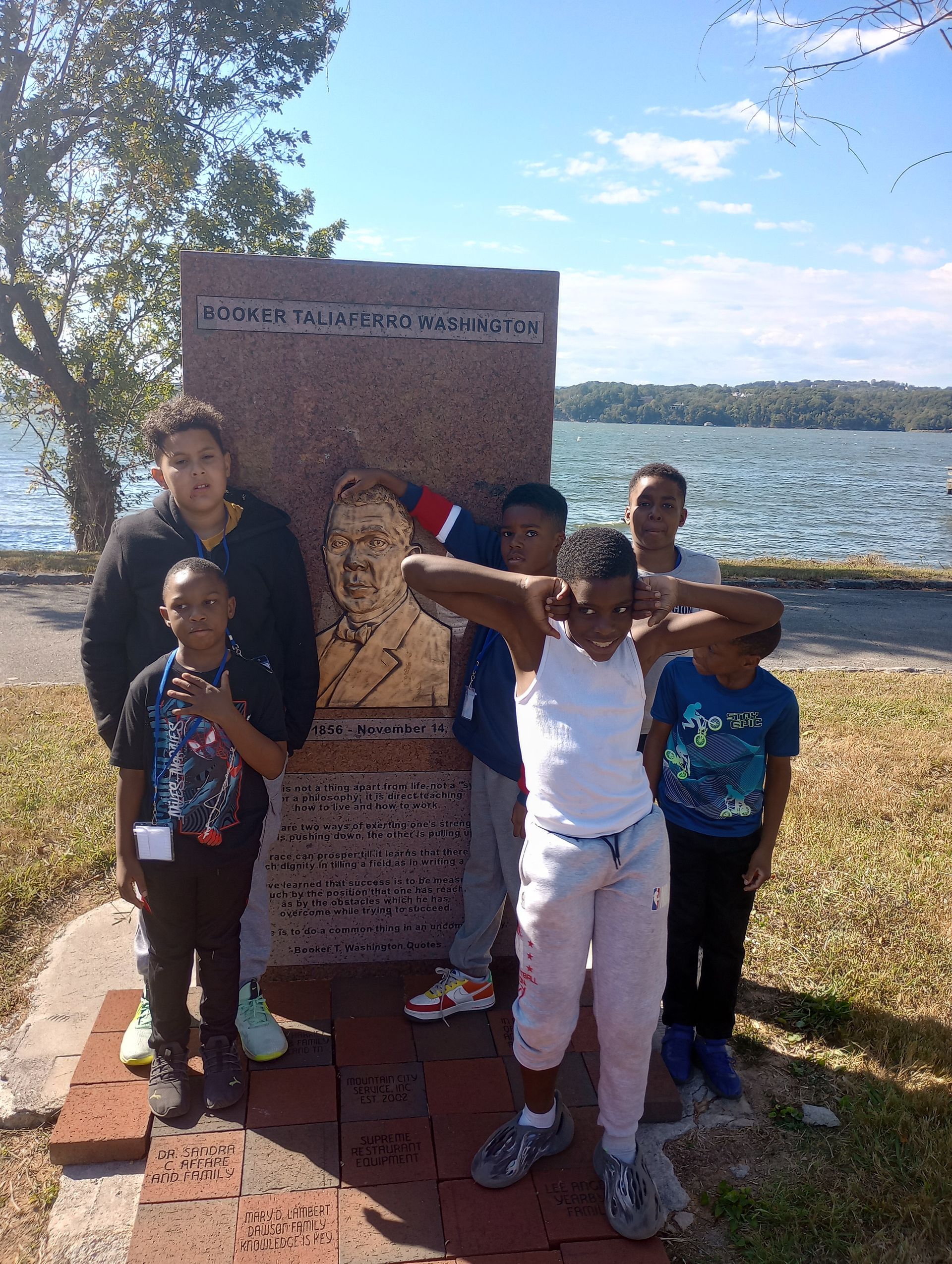 A group of children pose by a monument with a bust of Martin Luther King Jr. by a lake, blue sky overhead.