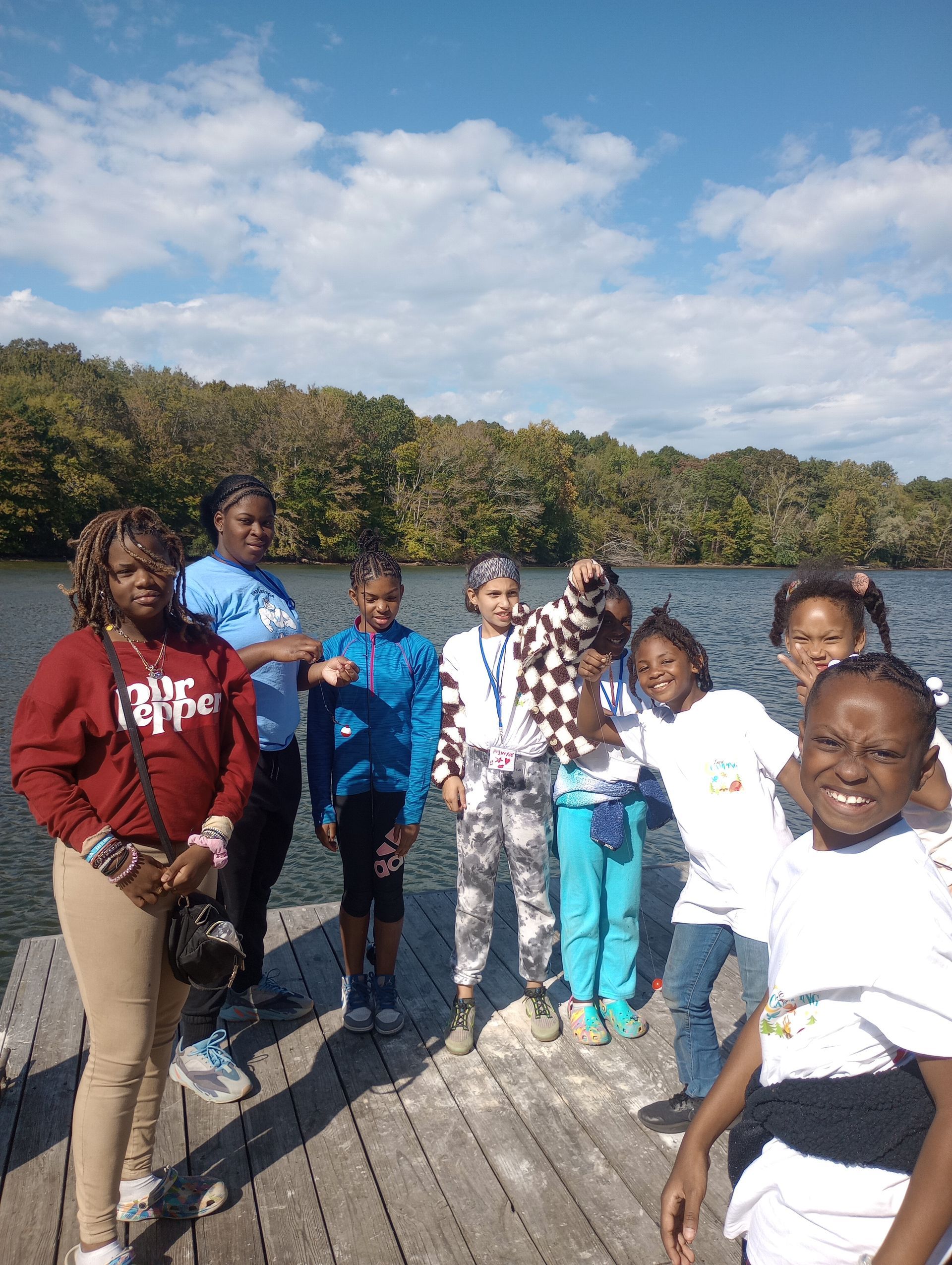 Group of children and an adult pose on a wooden dock by a lake on a sunny day. They smile and wear various colorful outfits.