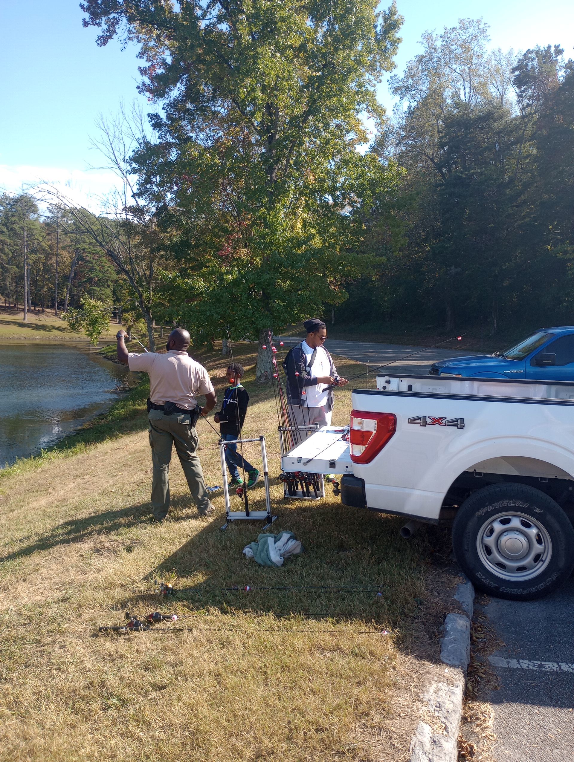 Man in uniform gestures near pond with two people and a truck parked on grassy area.