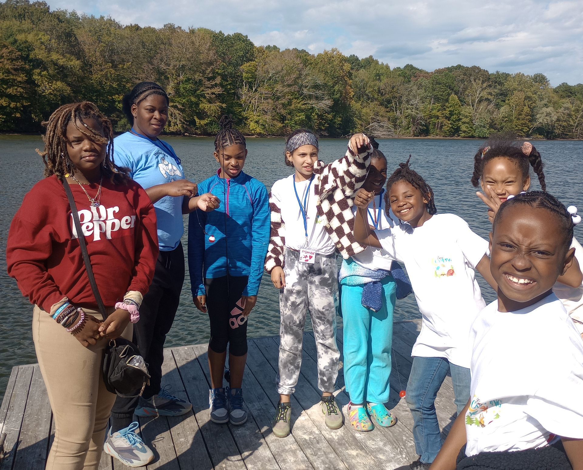 A group of children and an adult pose on a wooden dock by a lake, smiling. Trees and a cloudy sky are in the background.