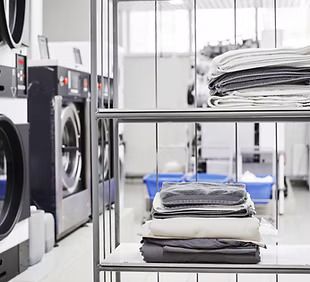 Laundry room interior. Folded clothes on shelves in the foreground. Washing machines and other equipment in the background.