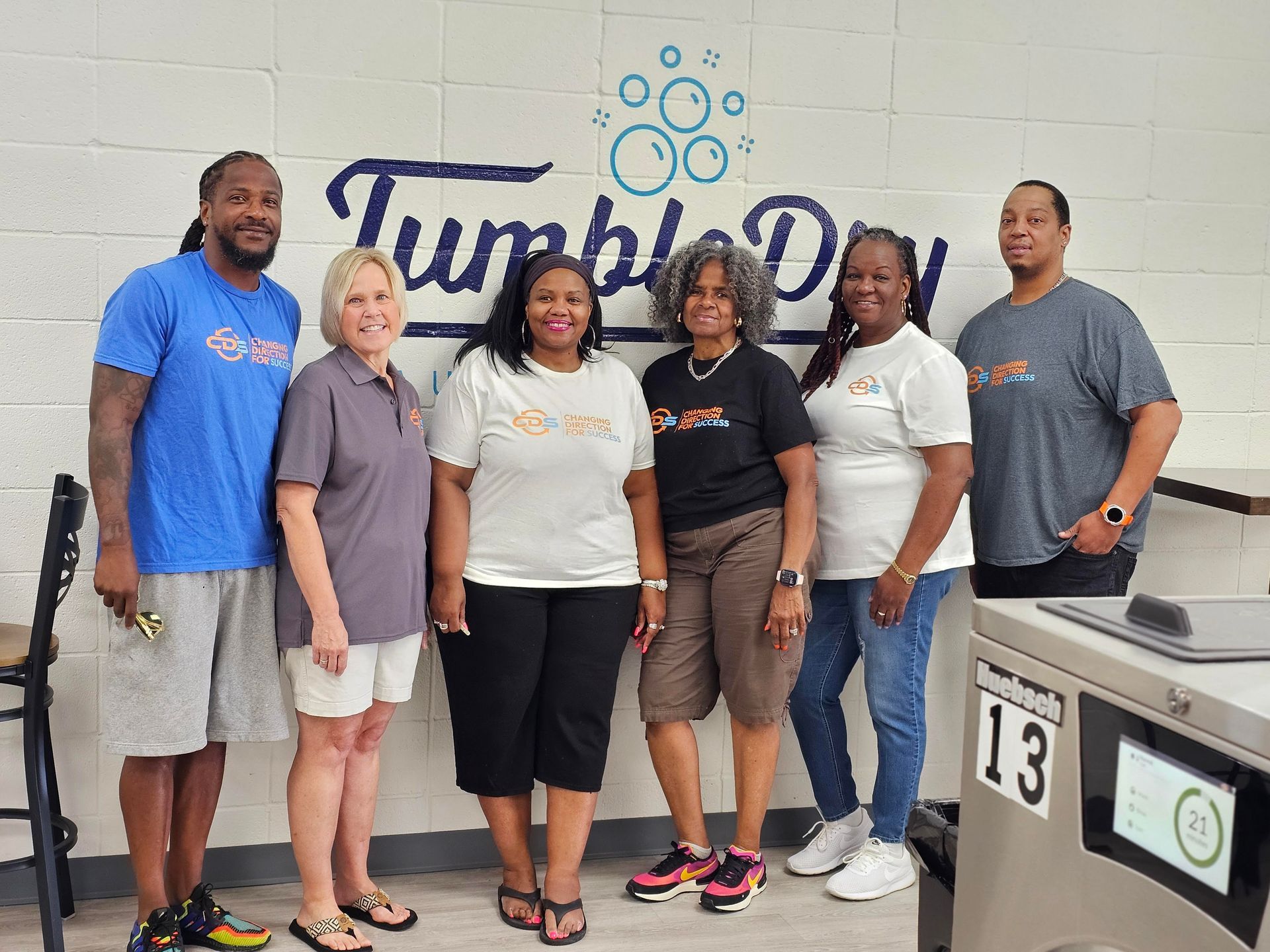 Laundry room interior. Folded clothes on shelves in the foreground. Washing machines and other equipment in the background.