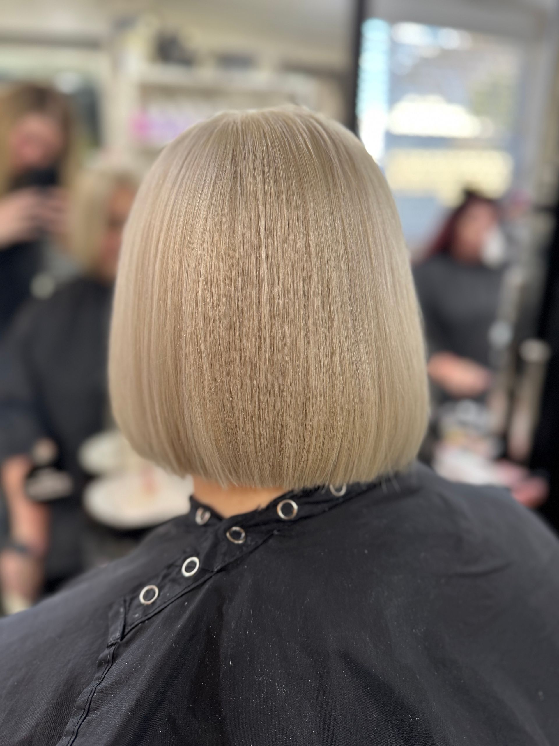 Woman with long, wavy blonde hair styled in a salon, facing a mirror. A stylist is behind her.