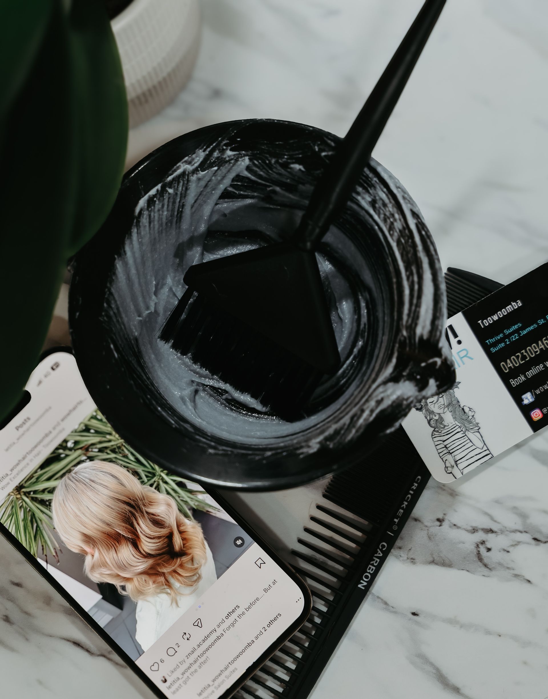 Hair dye bowl with applicator, phone showing hairstyle, combs on marble surface.