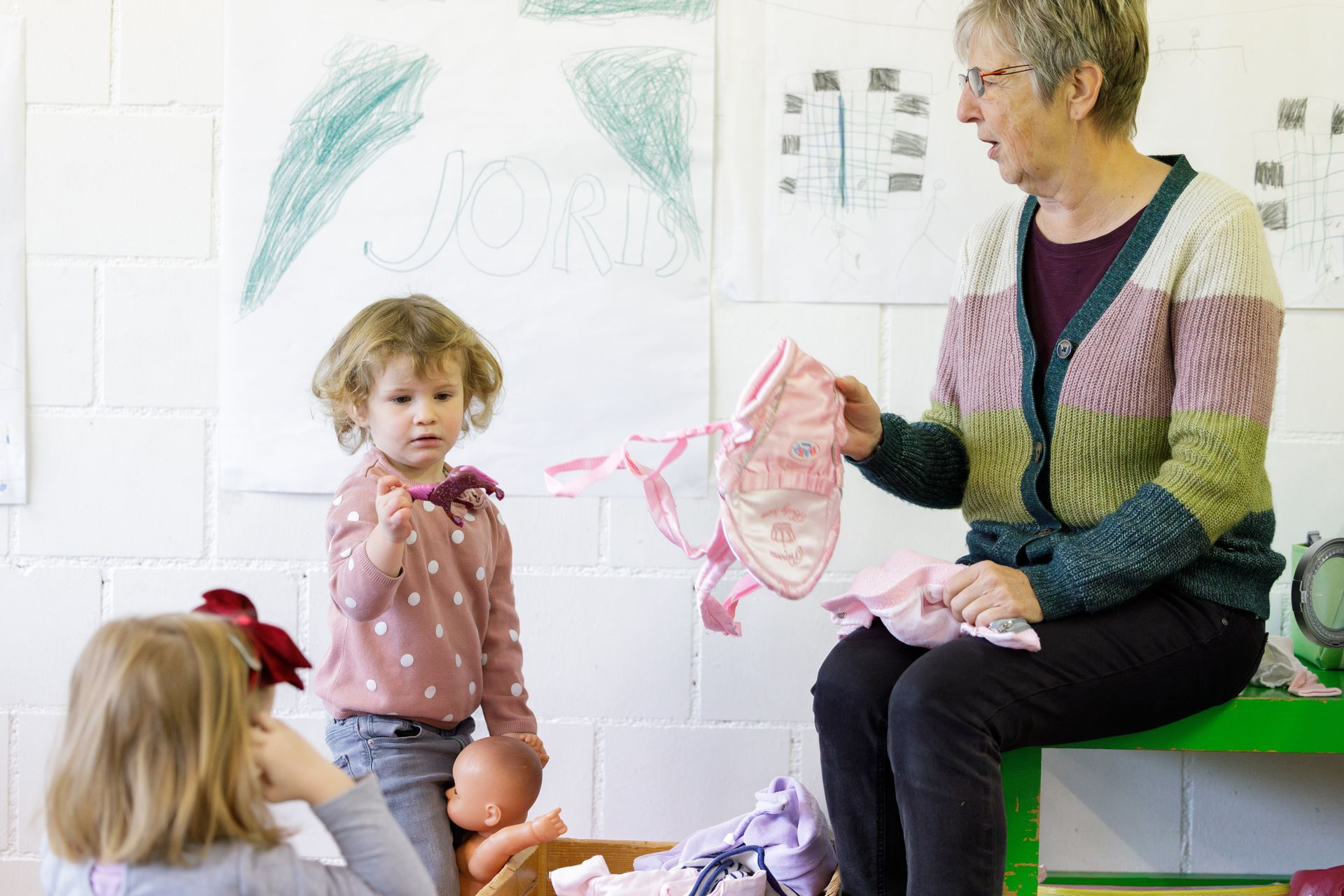 Eine Frau zeigt Kindern in einem Klassenzimmer Puppenkleider, ein Mädchen hält ein kleines Spielzeug.