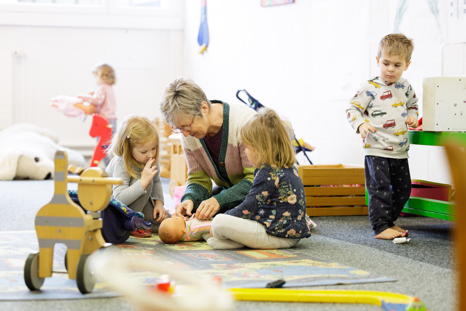 Kinder und ein Erwachsener spielen mit Puppen, im Hintergrund spielen ein paar andere Kinder.