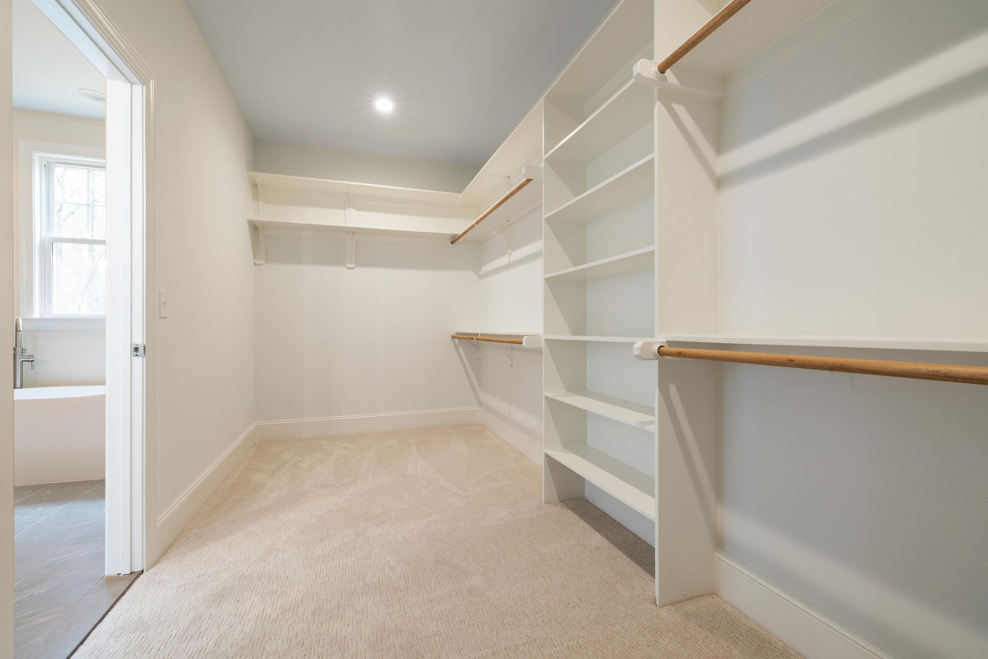 A walk in closet with lots of shelves and a bathtub in the background.