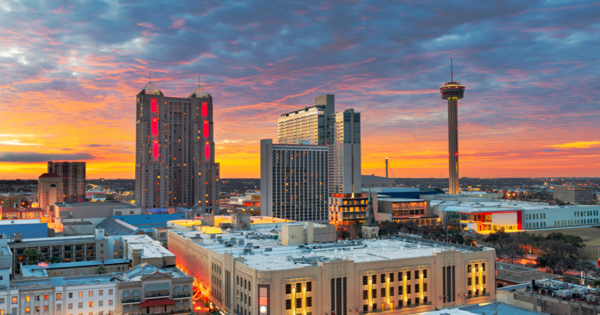 San Antonio skyline at sunset; prominent Tower of the Americas, colorful sky, and city buildings.