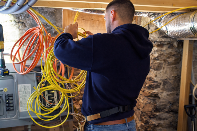 An electrician works with yellow and orange wiring at an electrical panel in a stone-walled basement.