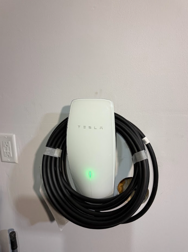 A white Tesla wall connector mounted on a wall with a coiled black charging cable and a solid green indicator light.