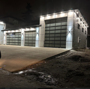 A modern detached three-car garage at night, illuminated by exterior wall and recessed soffit lights.