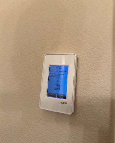 A white wall-mounted thermostat with a digital touchscreen display, shown at eye level on a beige wall.