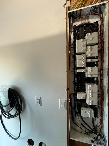 An open residential electrical breaker panel mounted on a white wall next to a wall-mounted EV charging cable.