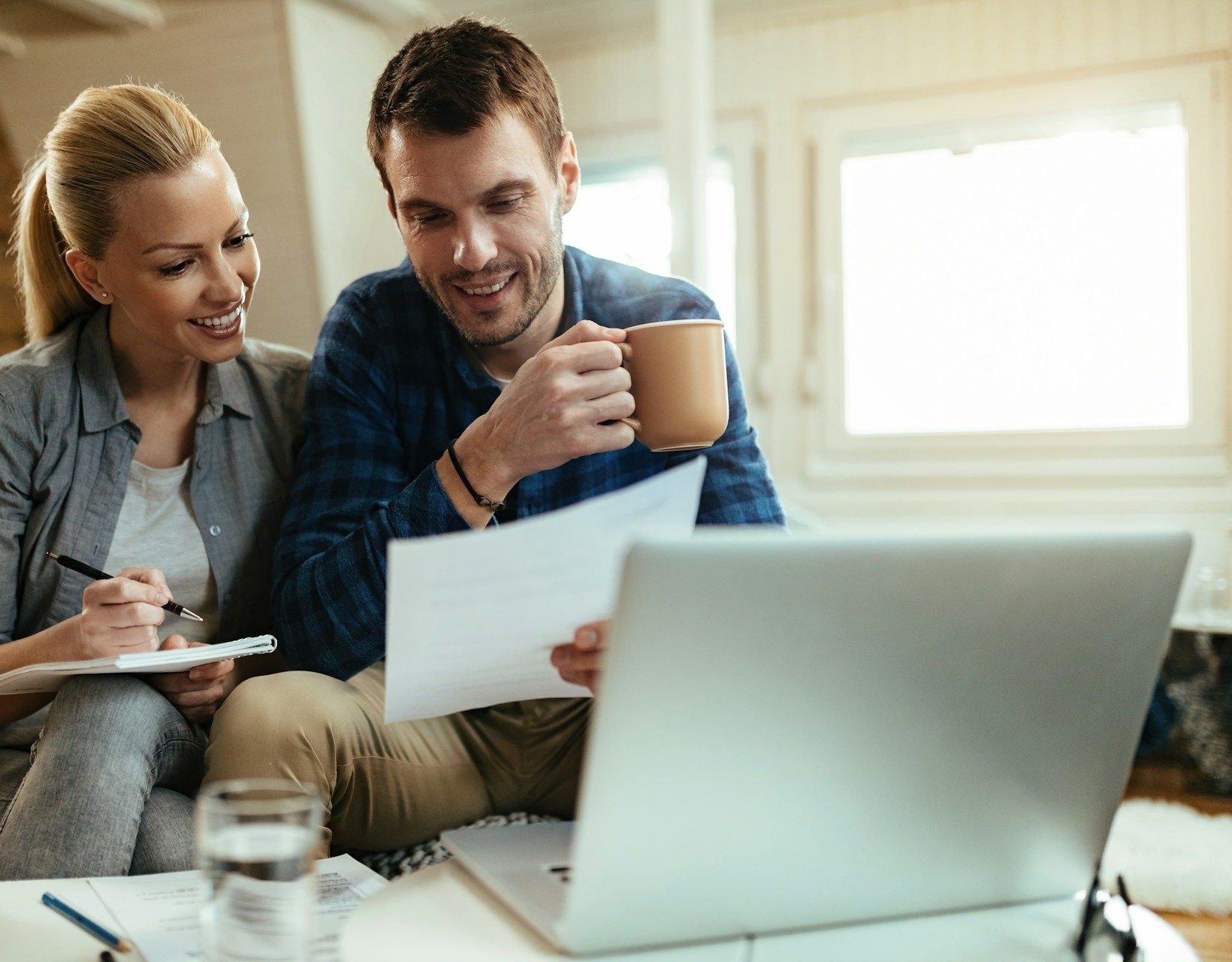 A person holding a coffee mug and another person writing on paper look at a laptop in a bright, modern living space.