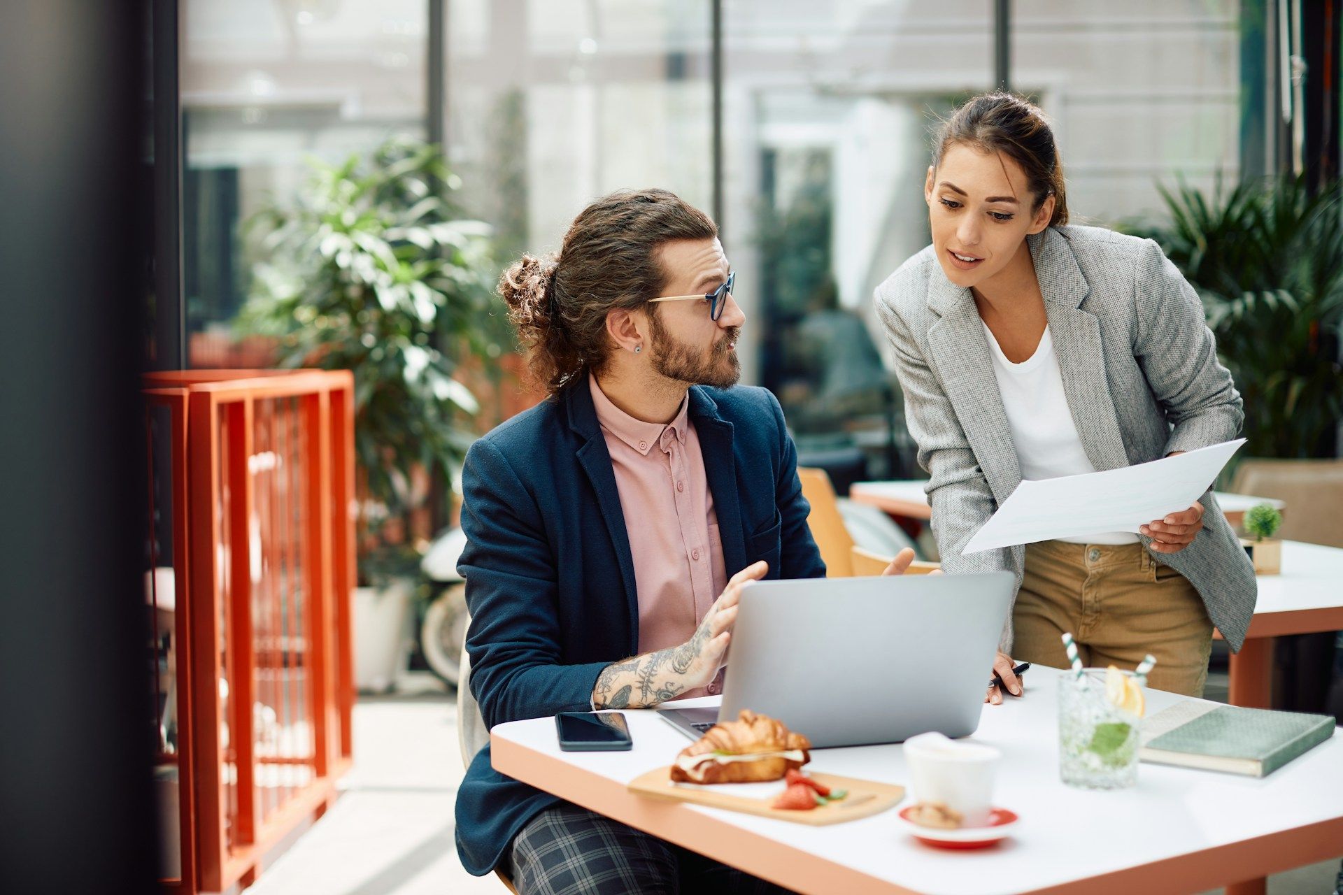 Woman in glasses and blazer types on laptop at a wooden table, notebook and phone nearby, with flowers in the background.