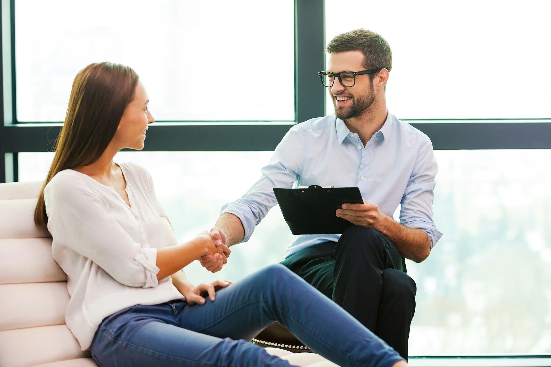 A smiling person with glasses holding a clipboard shakes hands with a seated person in a bright office by a window.