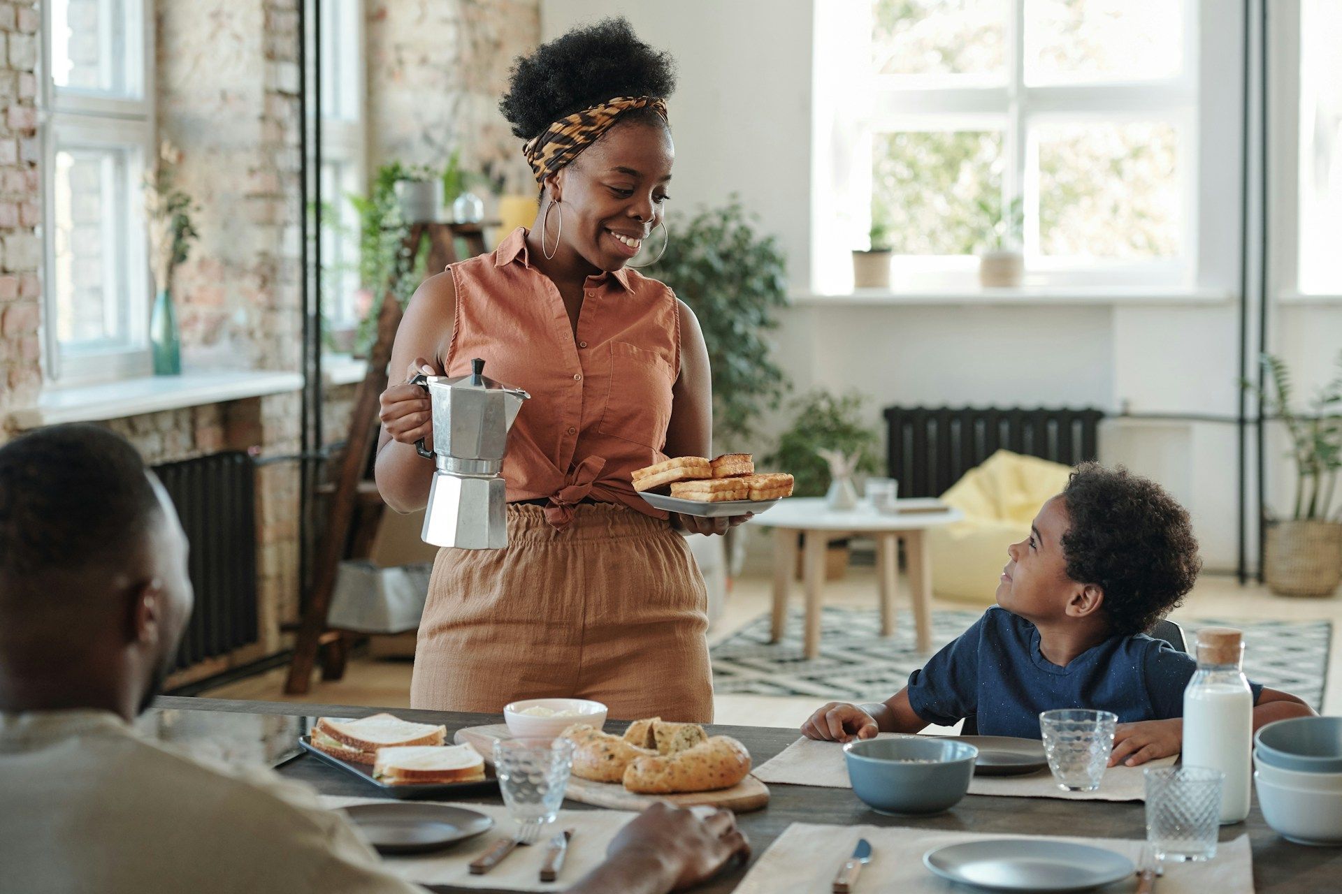 A person holds a coffee maker and a plate of food at a breakfast table while interacting with a young child.
