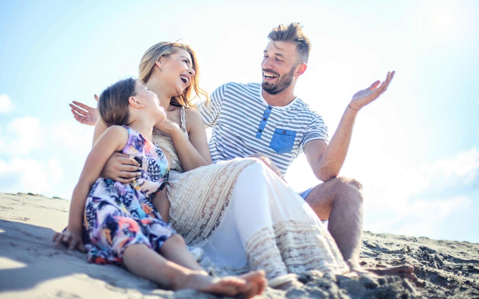 A family laughing together while sitting on a sandy beach under a bright, sunny sky.
