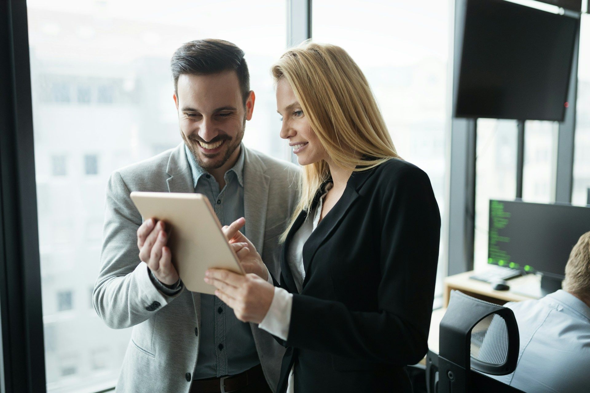 Two professionals in business attire stand by a window, smiling while reviewing information on a digital tablet.