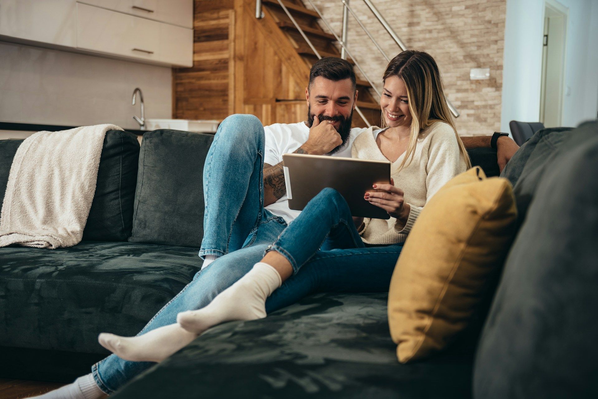 A couple sits on a dark sofa in a living room, looking at a tablet together and smiling.