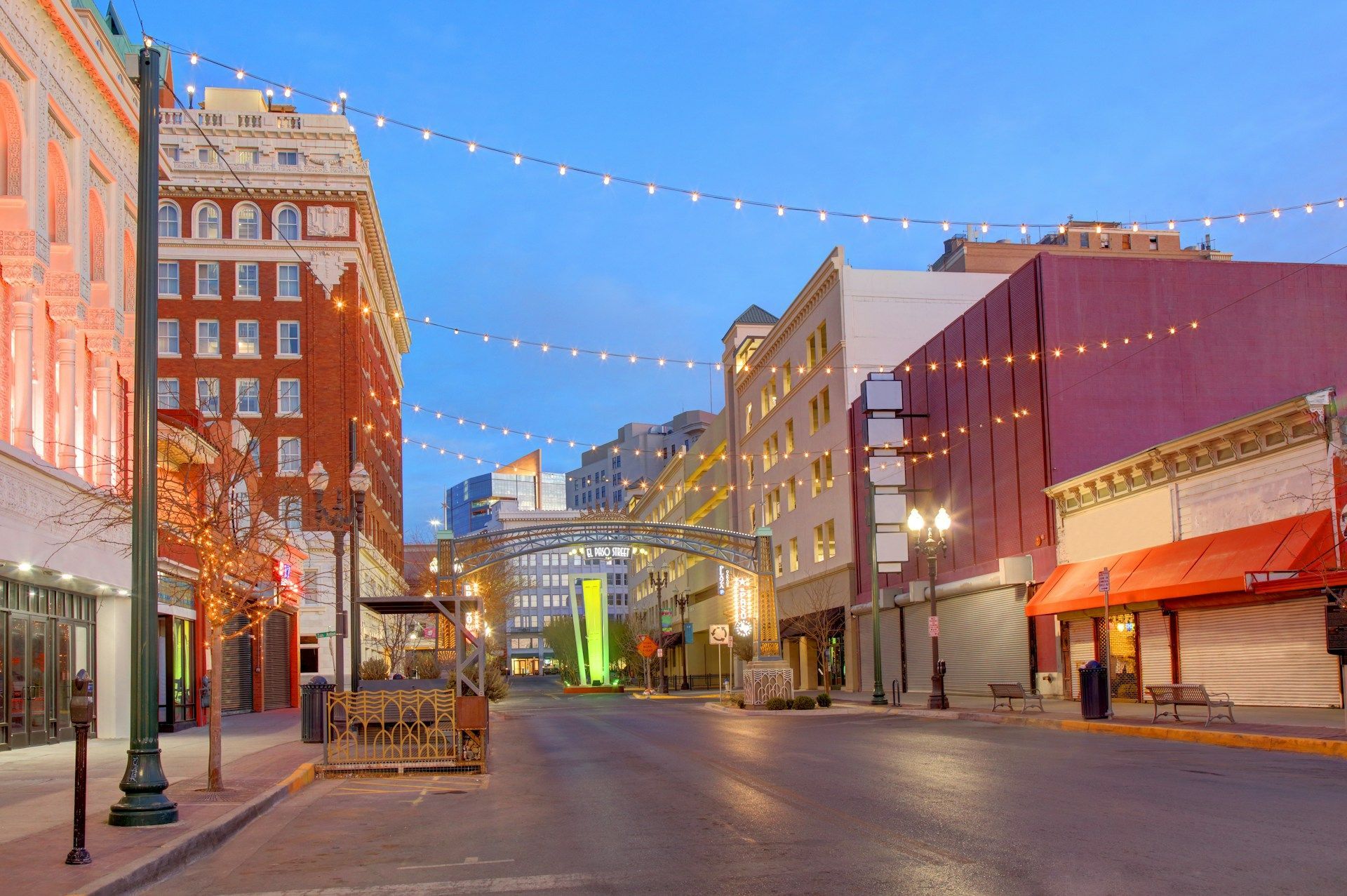 A street in downtown San Antonio at dusk, with string lights overhead, historic brick buildings, and an orange awning.