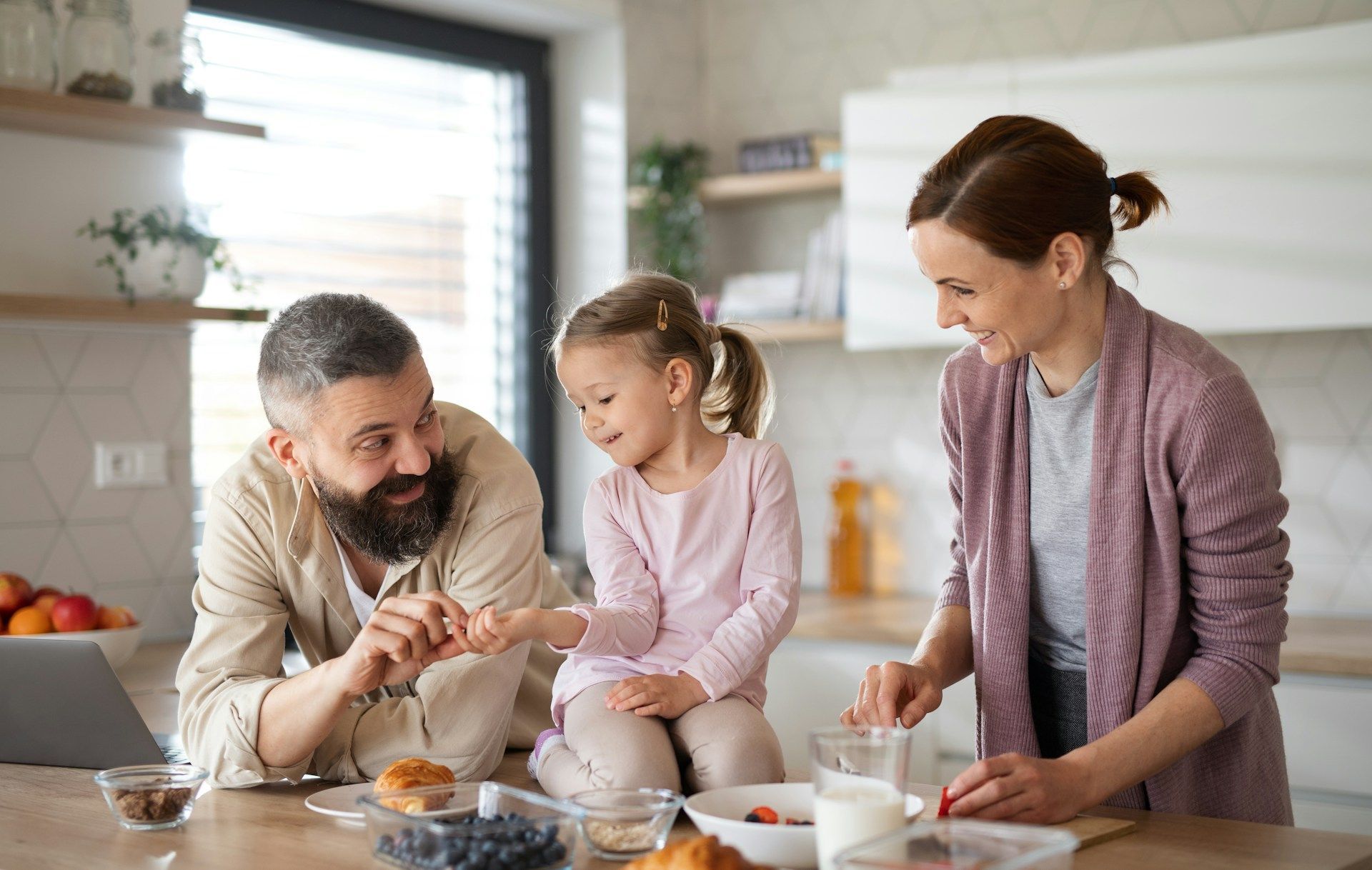 A family interacts warmly in a sunlit kitchen while preparing a meal together at a wooden table.