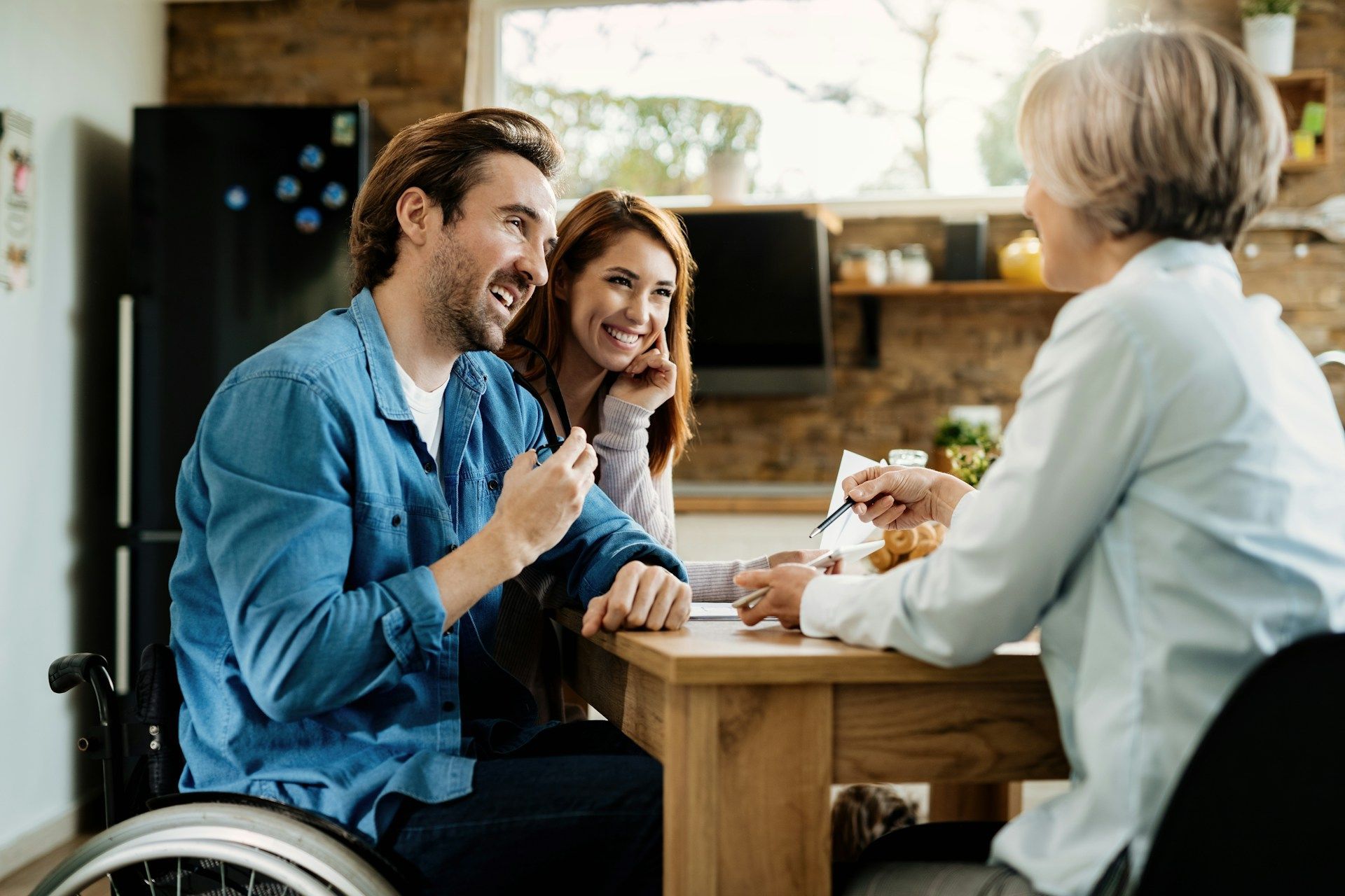 A person in a wheelchair and a woman talk with a third person at a wooden table in a kitchen setting.