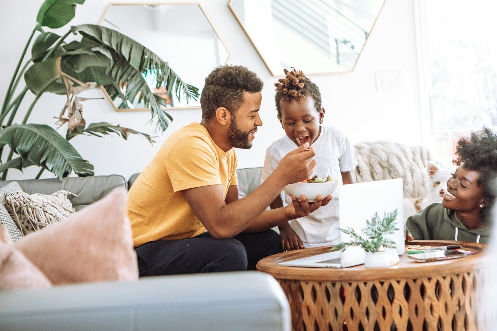 A person feeds a child from a bowl while sitting at a coffee table with another person in a sunlit living room.