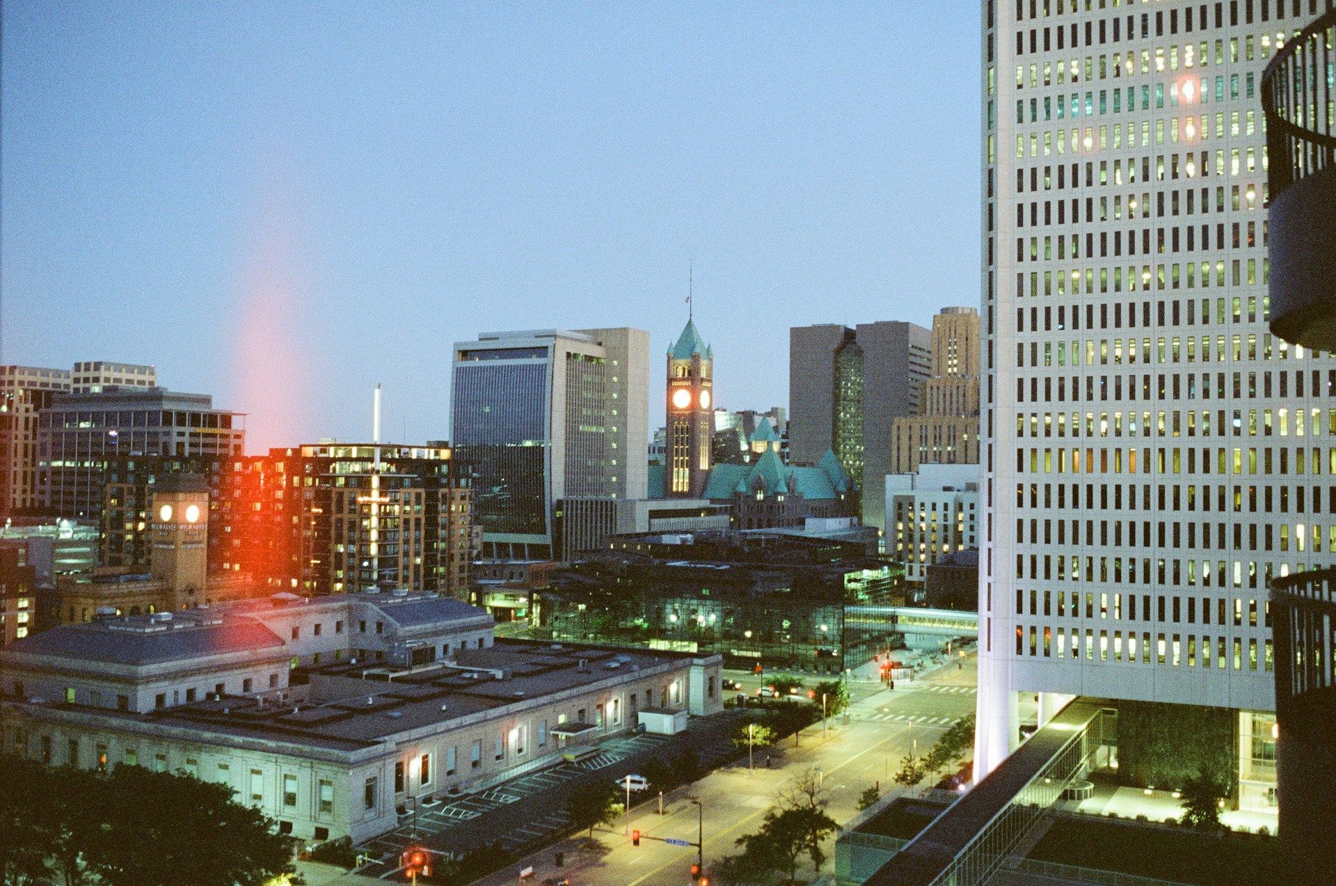 A twilight cityscape featuring modern high-rises, a historic stone building in the foreground, and a vertical light flare.