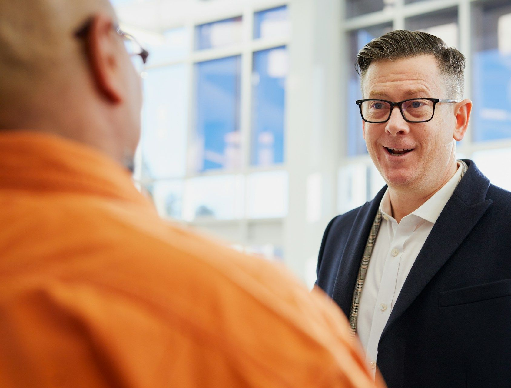 Two people in business attire talk in an office setting, with one facing the camera and smiling while speaking.