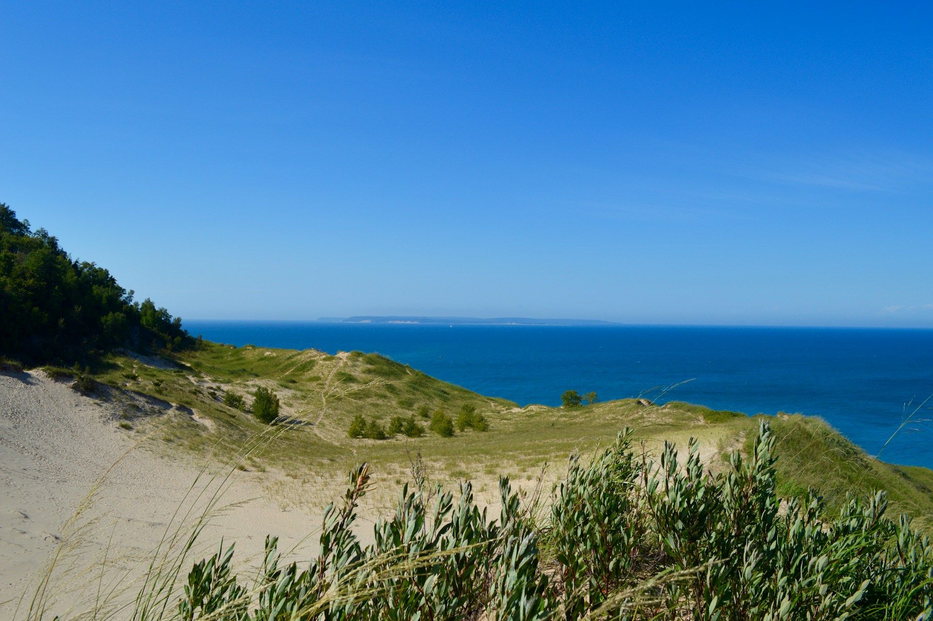 A scenic view of a grassy, sandy dune leading down to a deep blue lake under a clear, bright blue sky.
