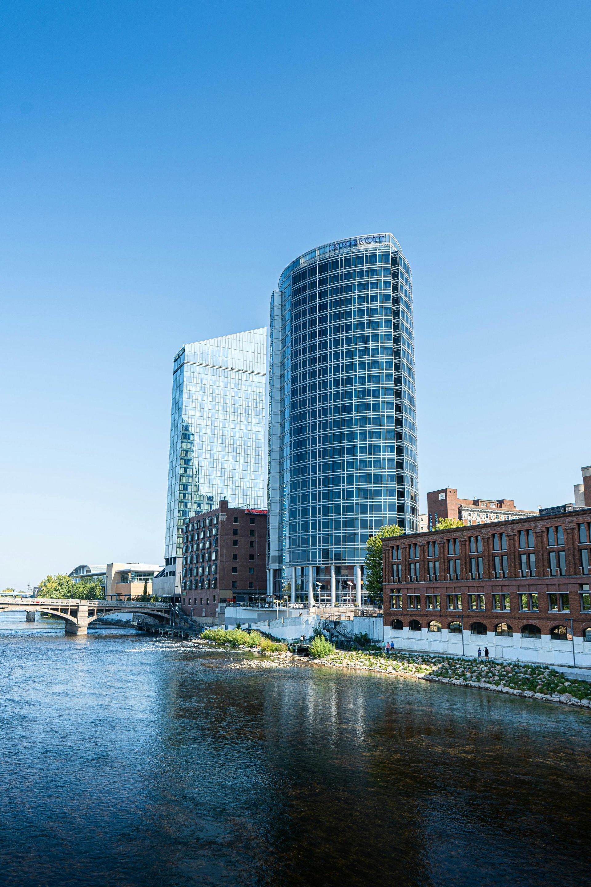 A modern glass skyscraper towers over older brick buildings beside a calm river under a clear blue sky.