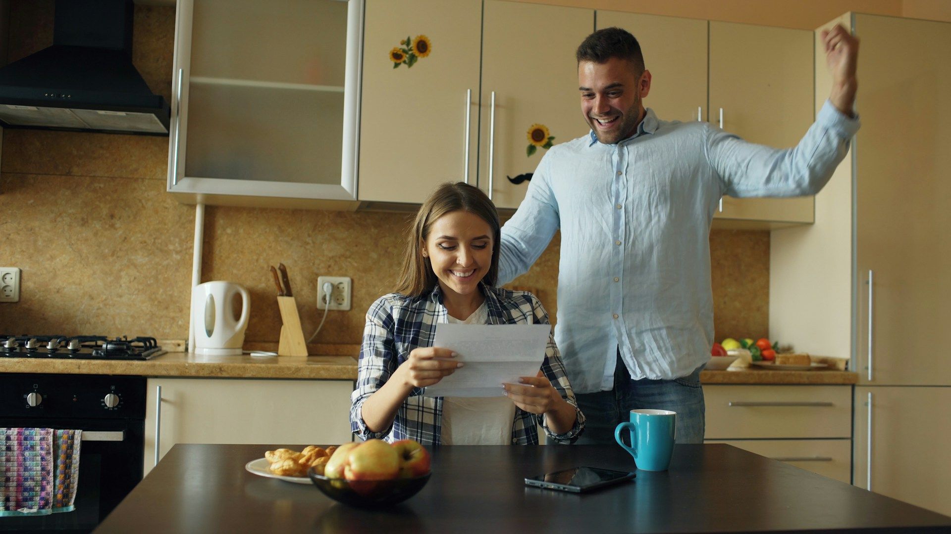 A person sits at a kitchen table reading a letter, while another person stands behind them celebrating with an arm raised.