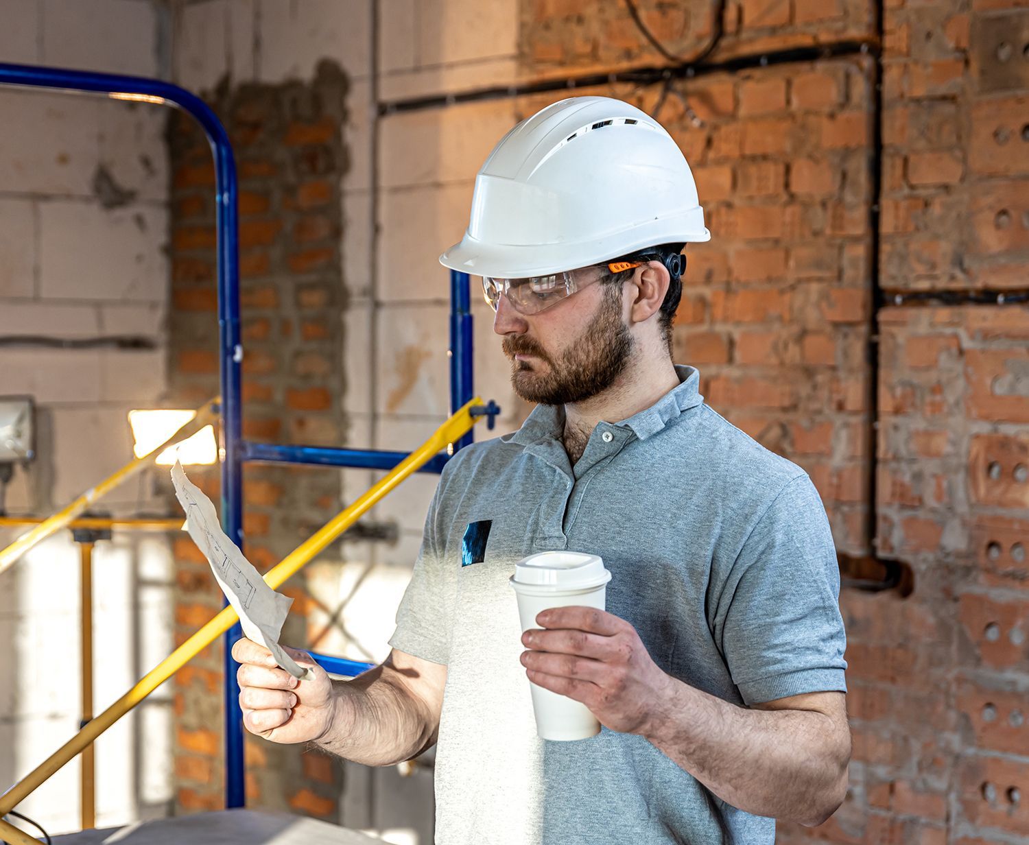 A man wearing a hard hat and glasses is holding a cup of coffee and looking at a piece of paper. A man wearing a hard hat and glasses is holding a cup of coffee and looking at a piece of paper.