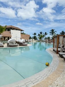 Luxury resort pool with turquoise water, palm trees, lounge chairs, and a blue sky with clouds