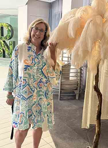 Woman standing indoors beside a large cream feather arrangement, wearing a colorful patterned dress.