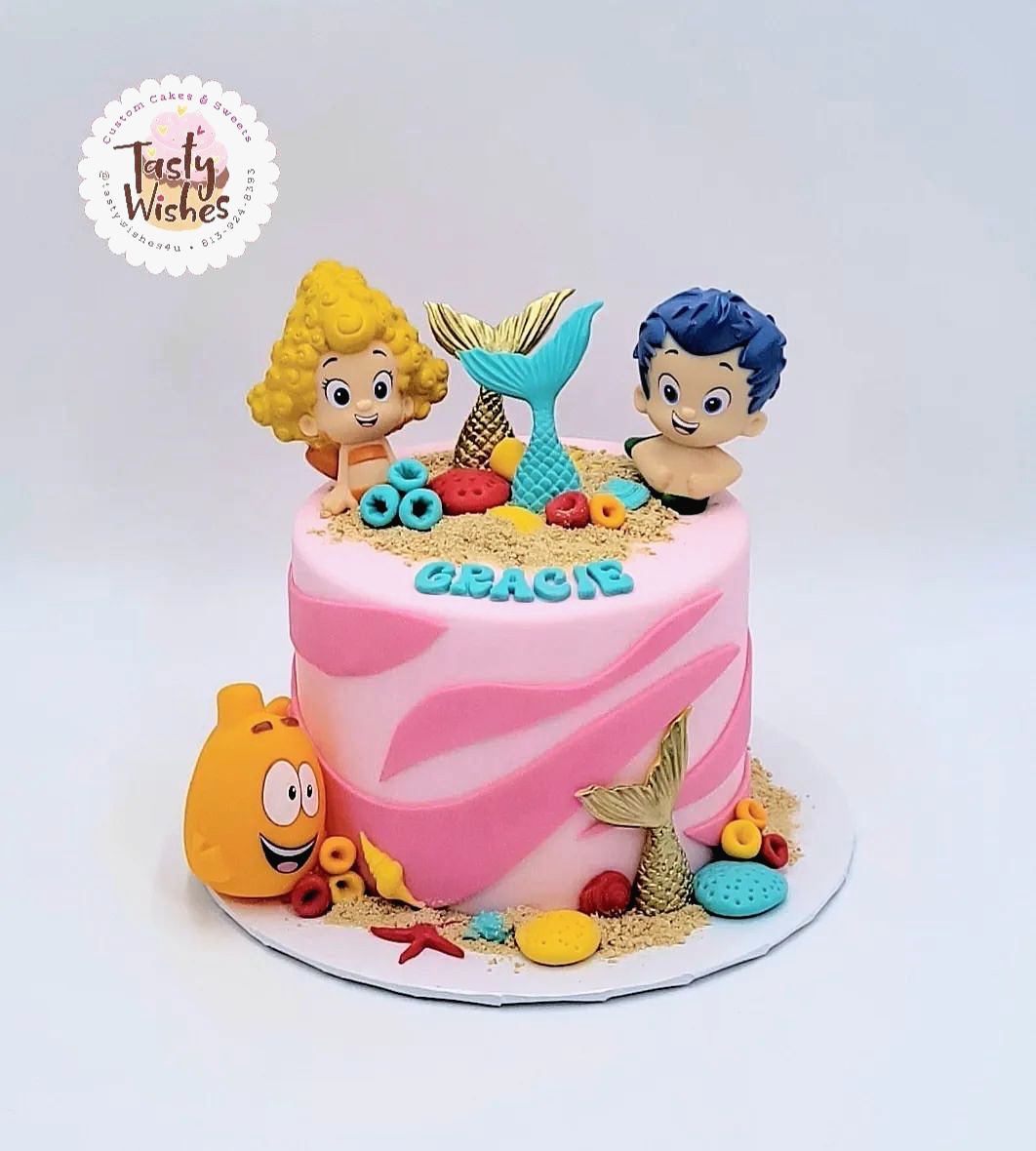 A decorated birthday cake with a light pink frosted base, topped with Bubble Guppies figures and
