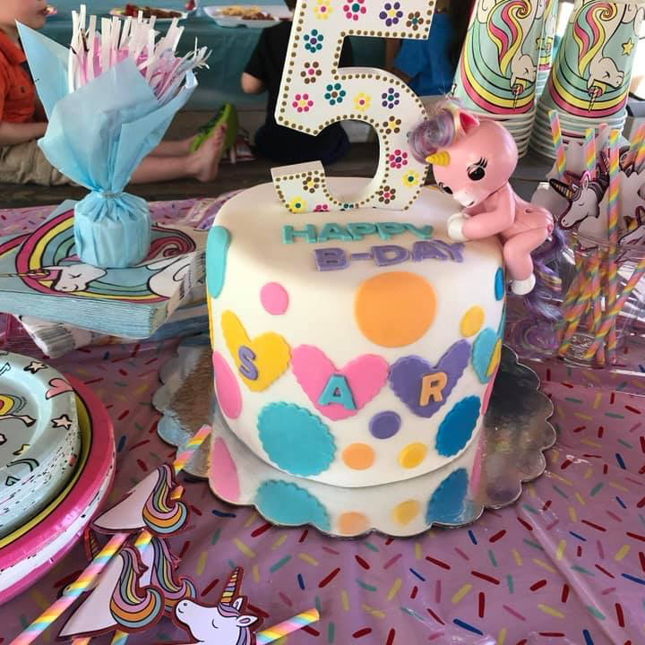 A 5th birthday cake decorated with colorful hearts and dots, featuring a unicorn figure and the name SAR on a party table.