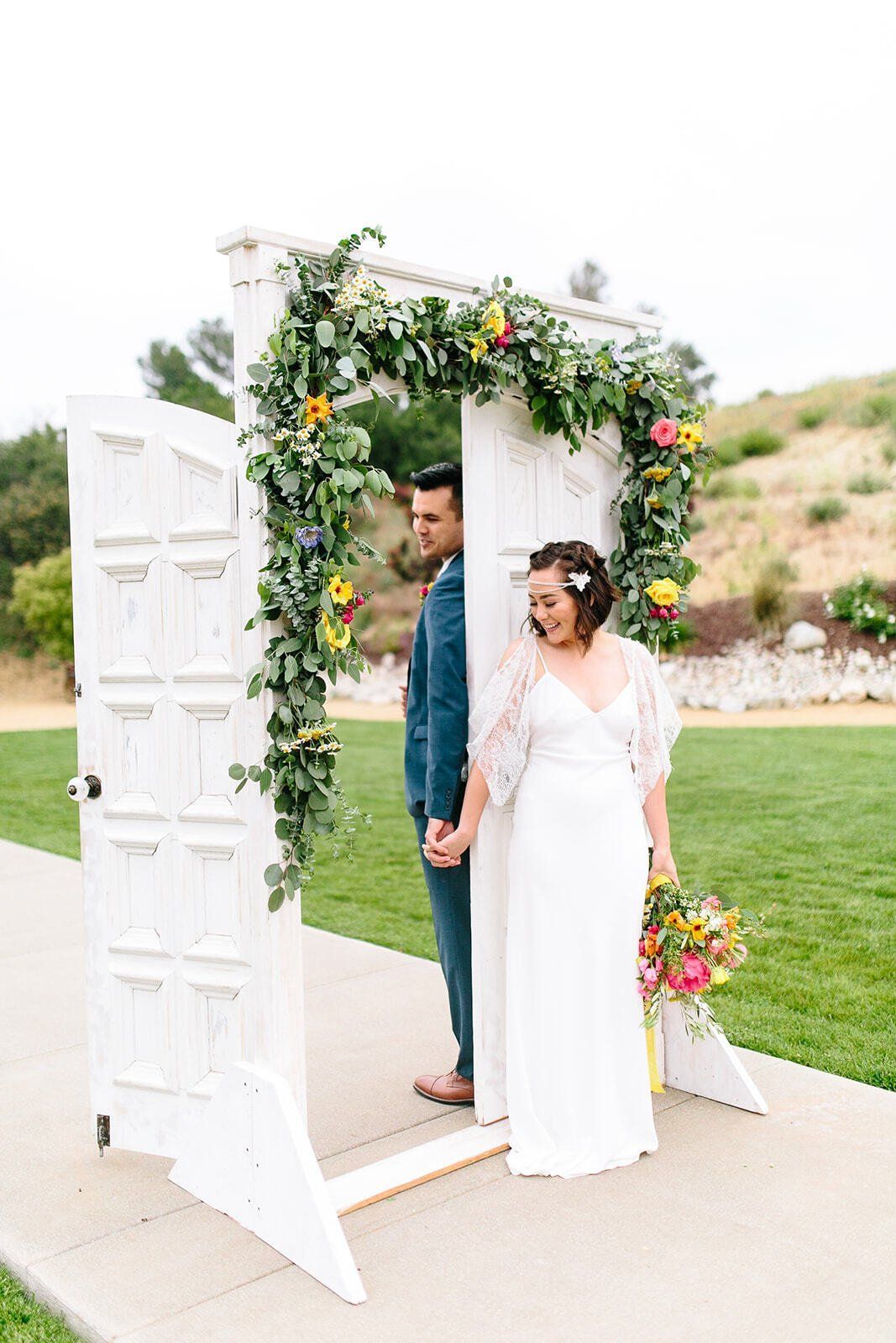 A bride and groom are standing in front of a white door decorated with flowers.