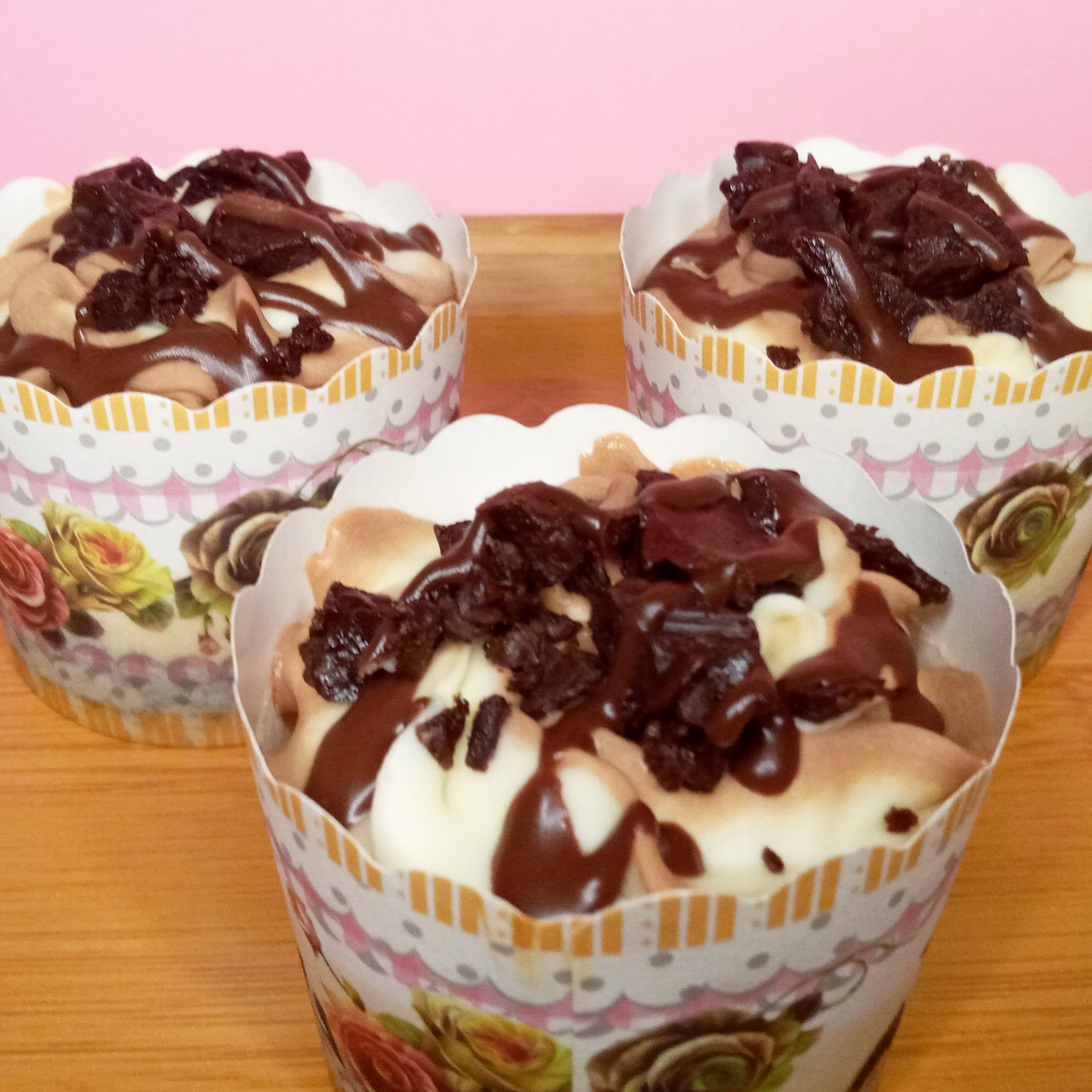 Three cupcakes are sitting on a wooden table with a pink background