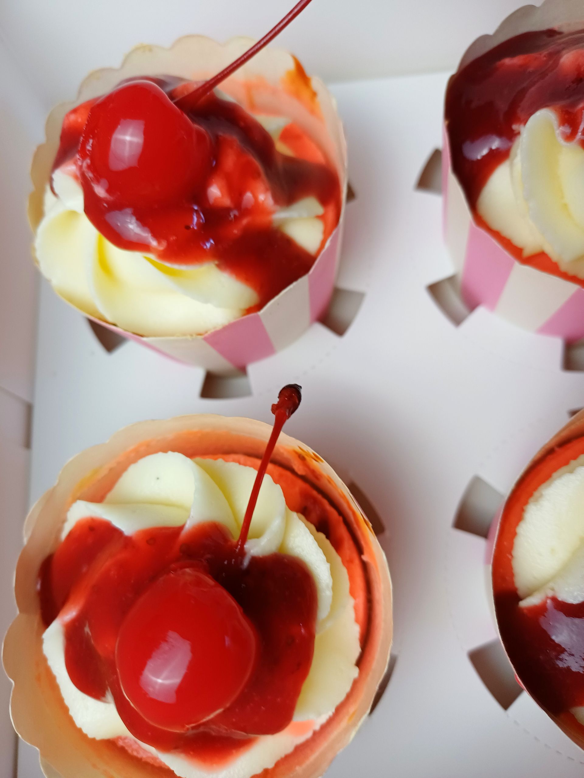 Four cupcakes topped with white frosting, red cherry sauce, and a maraschino cherry in a white box.